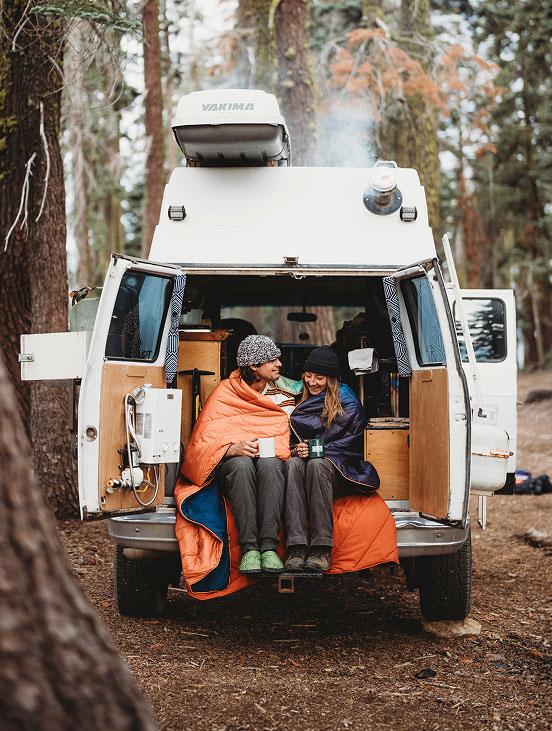 Couple wrapped in blankets sitting in a camper van in a forest.