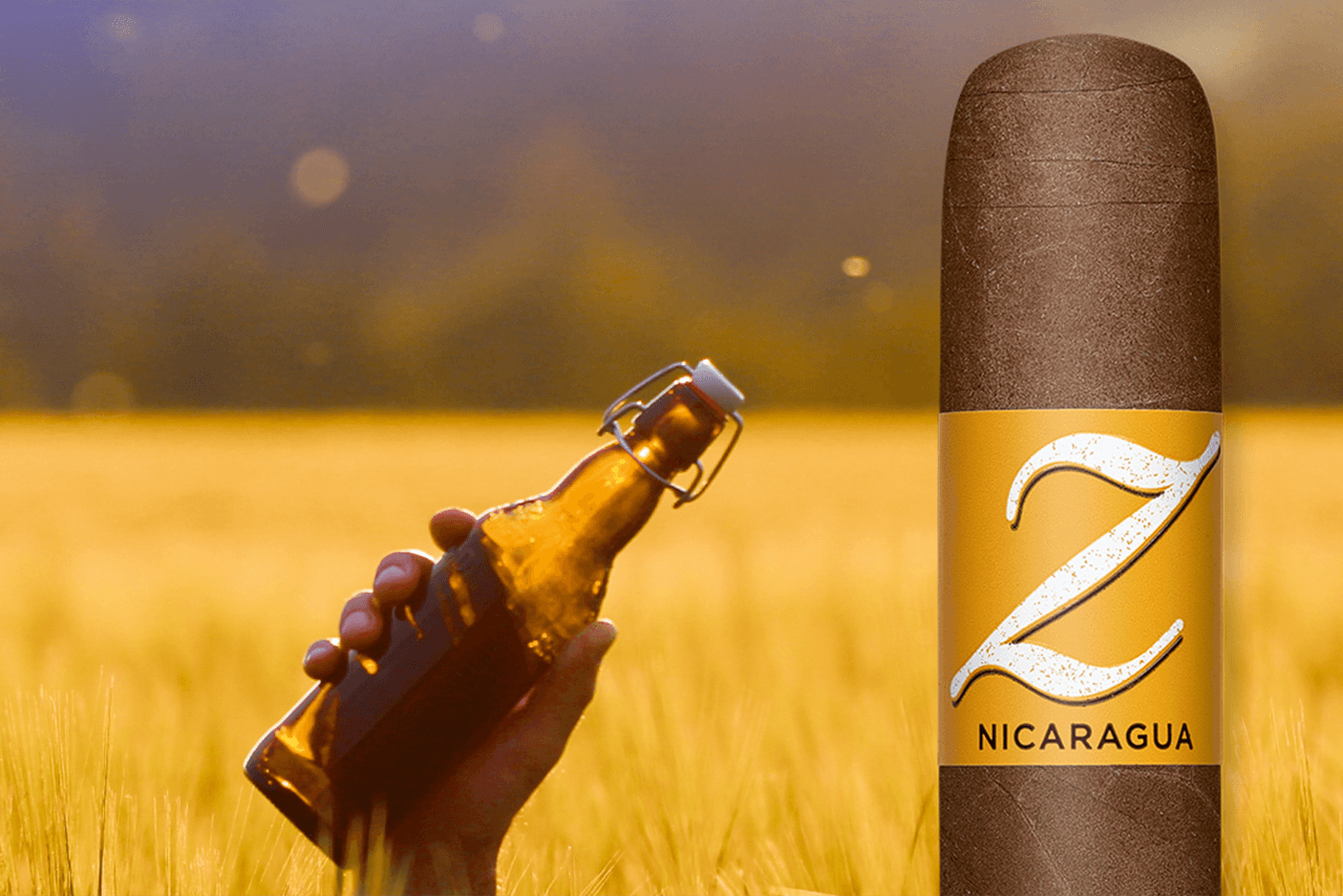 A Zino Nicaragua cigar in front of a background with a hand holding a beer bottle up in the air in a corn field. 