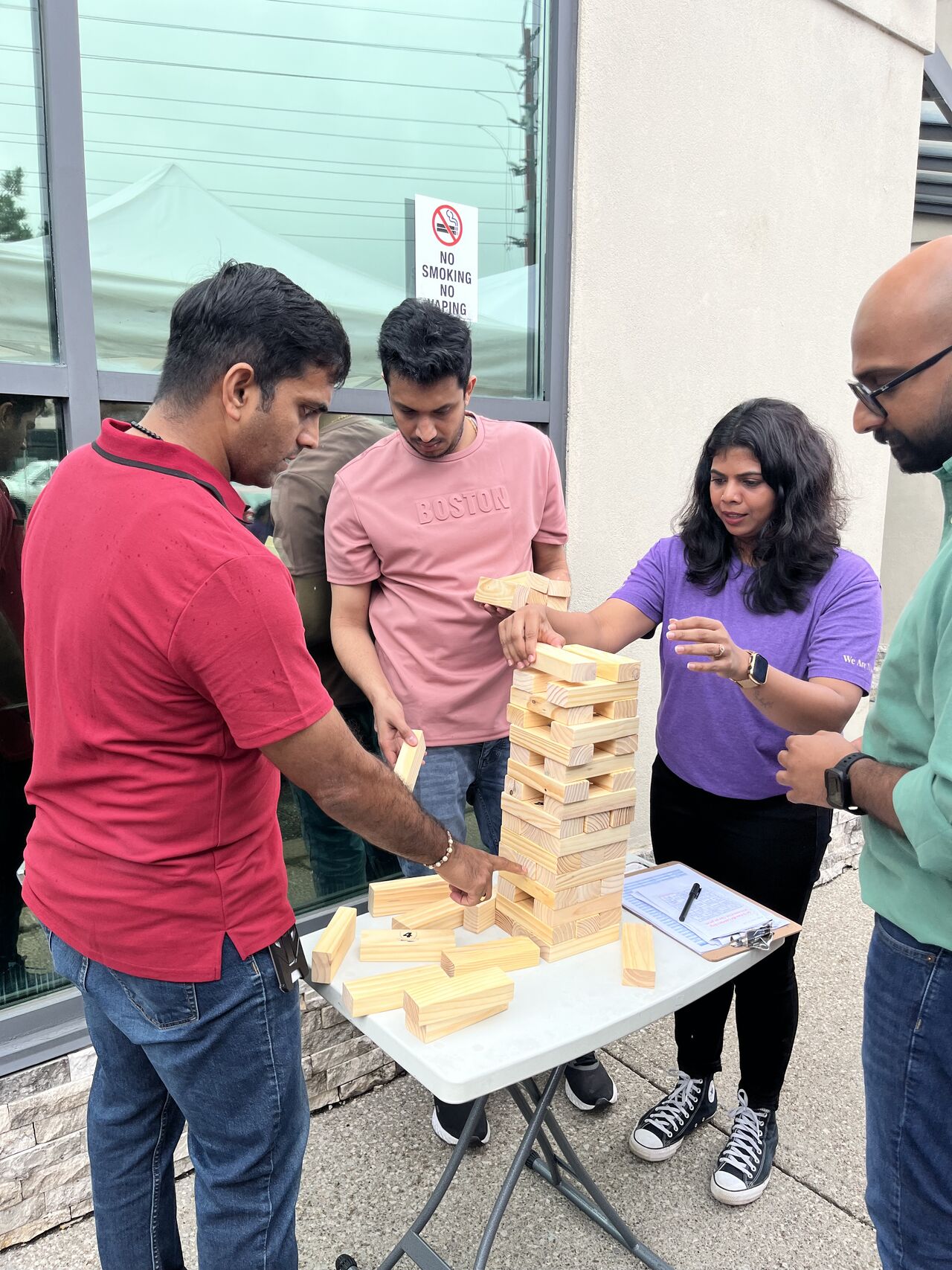Four people playing a block stacking game outdoors at a table.