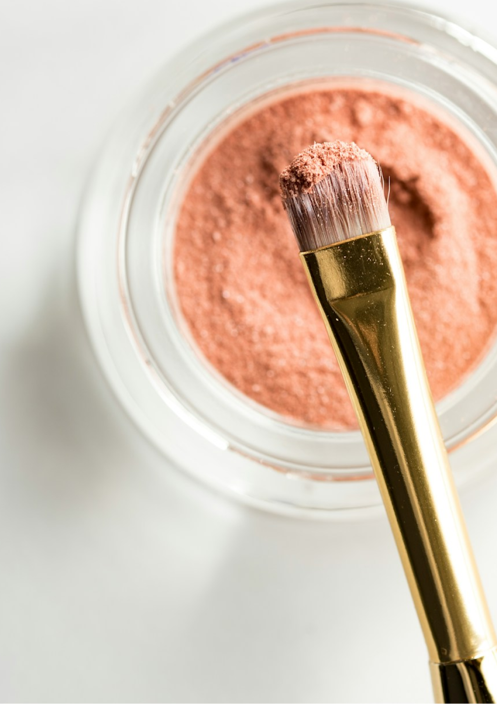 A glass pot containing makeup powder, with a gold-clad brush hovering over it, viewed top down.