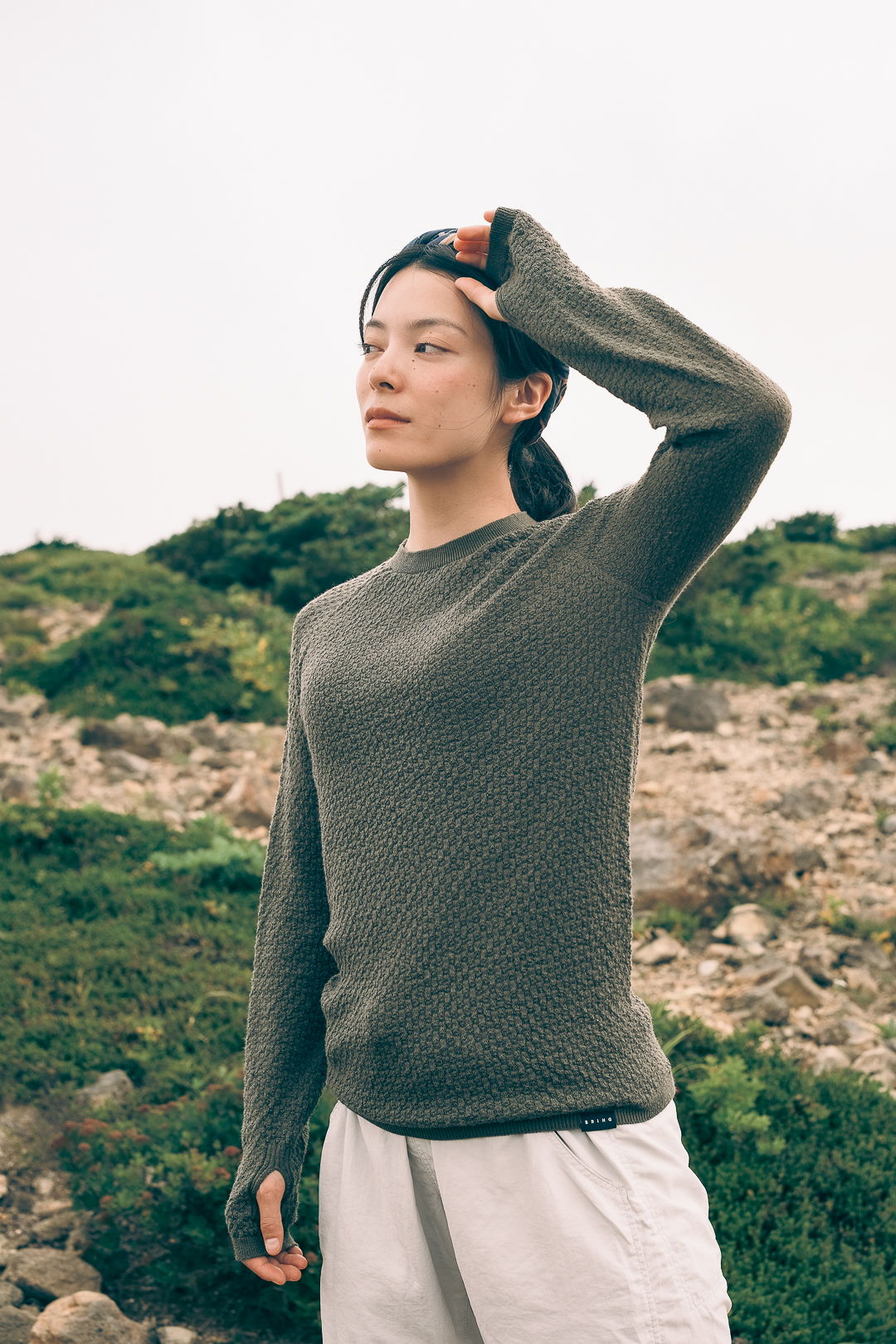 A woman in a green sweater stands outdoors, touching her hair.