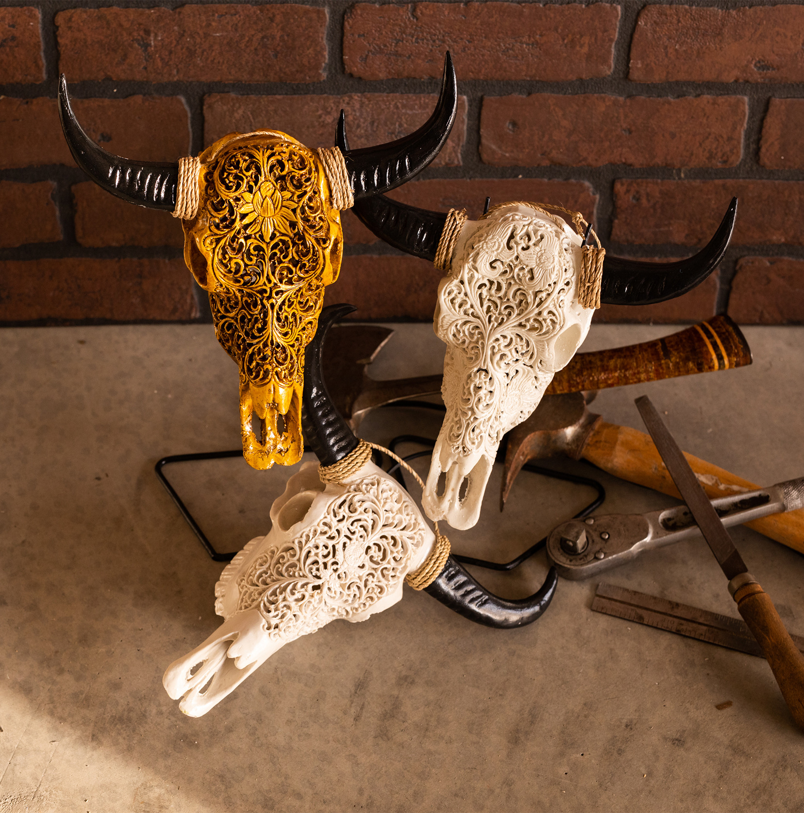 Three hand carved skulls are presented next to some tools in front of a brick wall. 