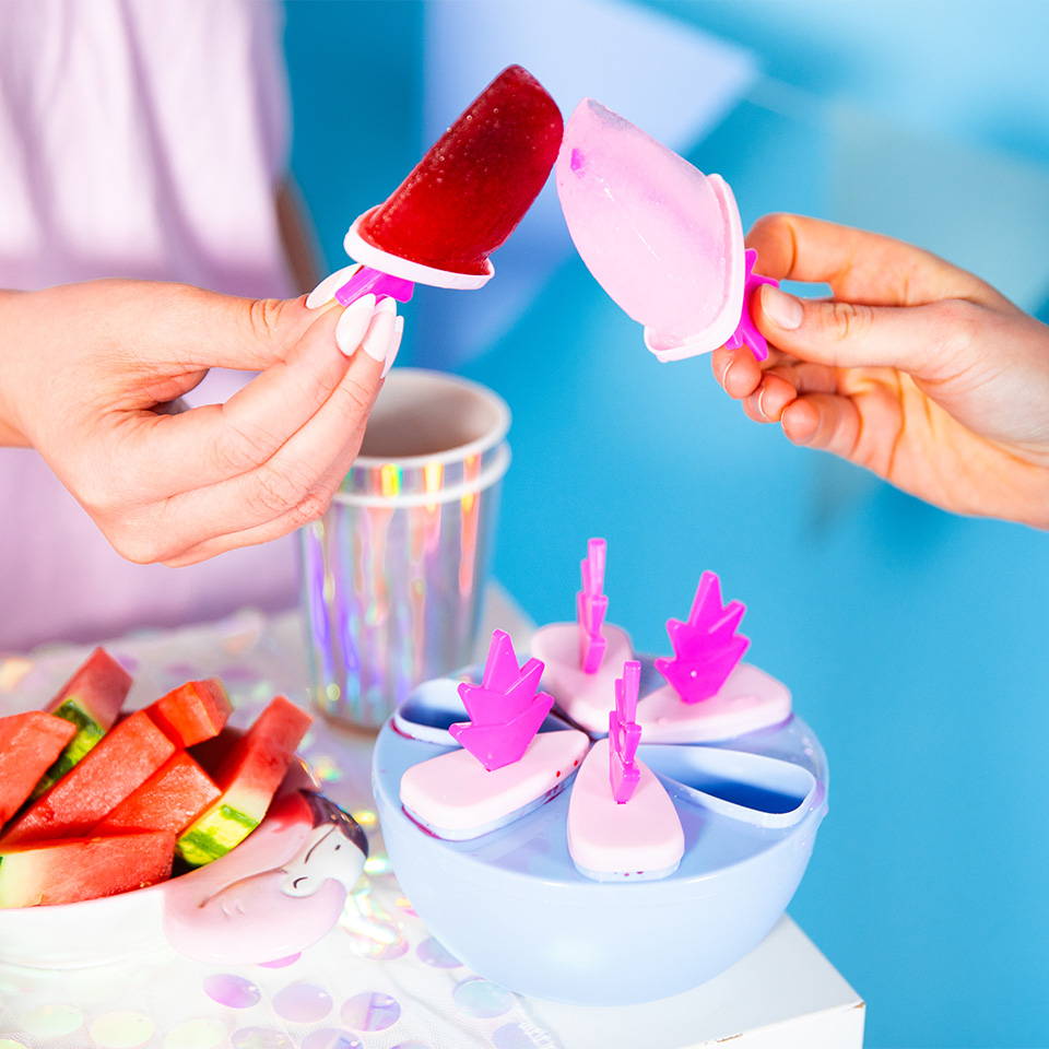 Hands holding flamingo-shaped popsicles near a flamingo popsicle mold set on a table with a tropical backdrop.
