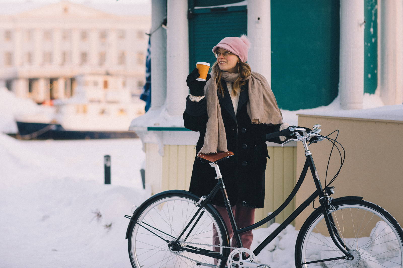 Woman outdoors in the snow wearing winter clothing and holding a coffee cup next to black step through city bike