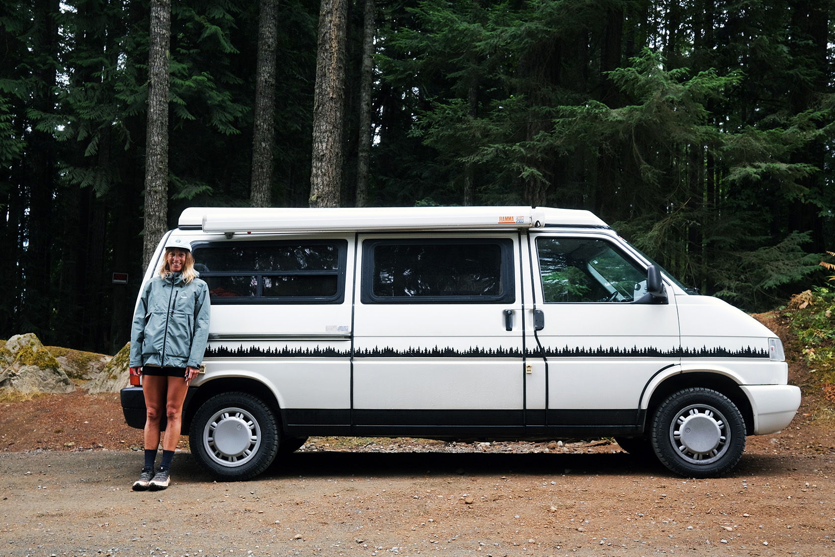 Person standing next to a white camper van in a forested area.