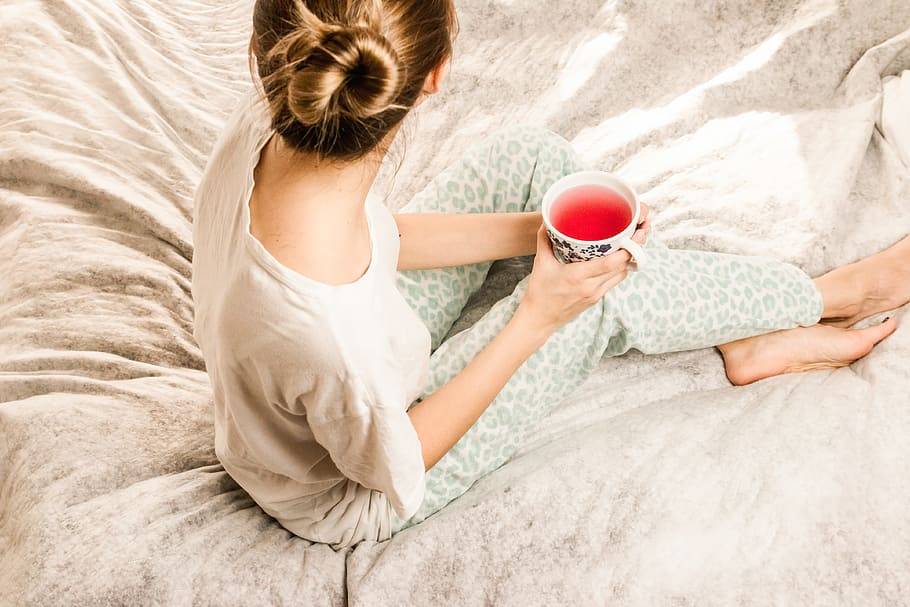 A young blonde woman with her hair in a bun sitting on a bed holding a mug of tea