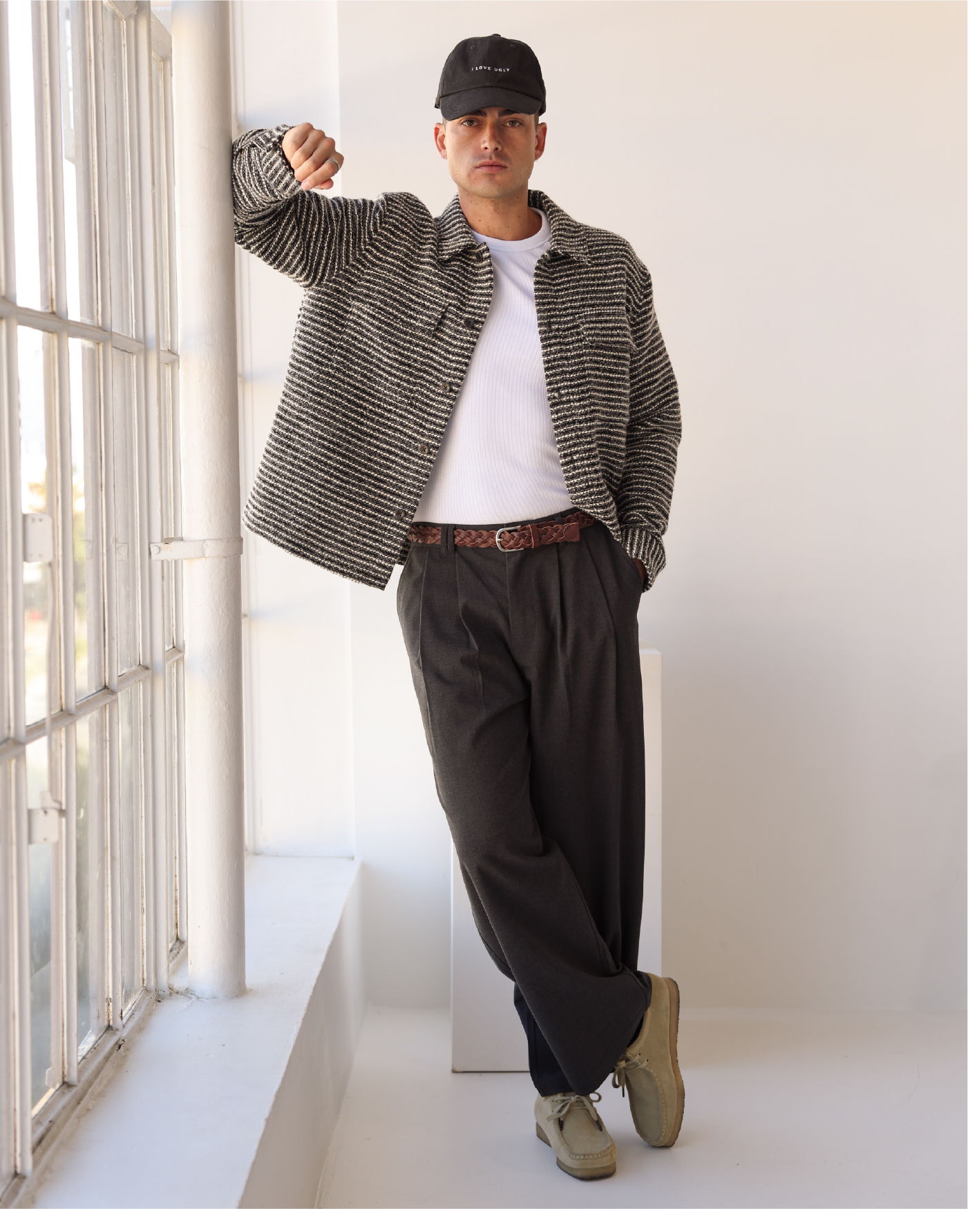 Person in casual outfit leaning against a wall near large windows.