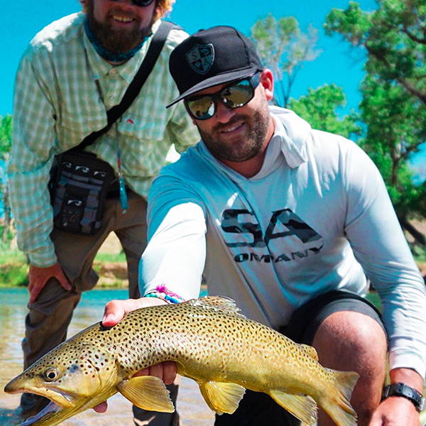 Ryan Copenhaver holding a fish up to the camera while wearing sunglasses, and SA Company hat and hooded performance shirt.