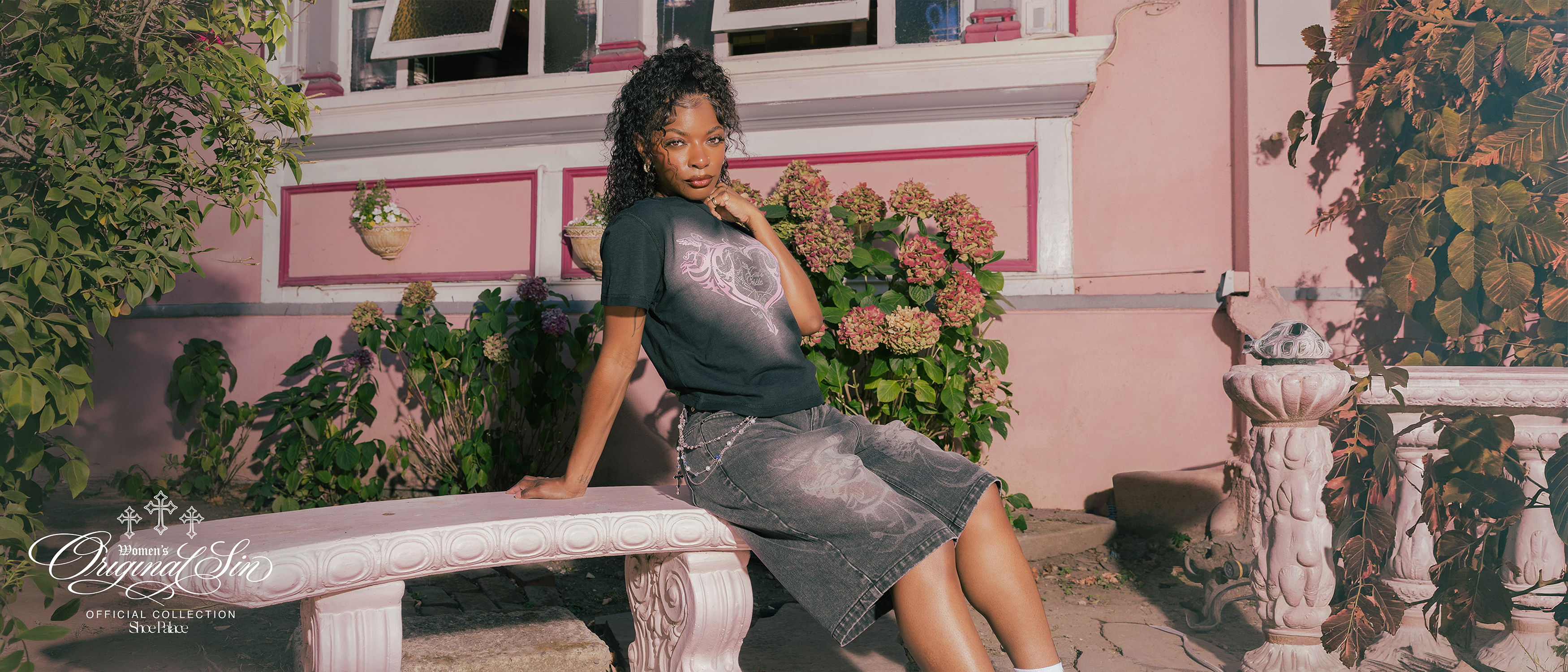 Person sitting on a bench with plants and a pink building in background.