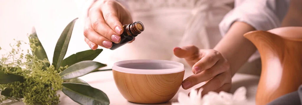 Person adding oil to a wooden bowl surrounded by herbs and a teapot.