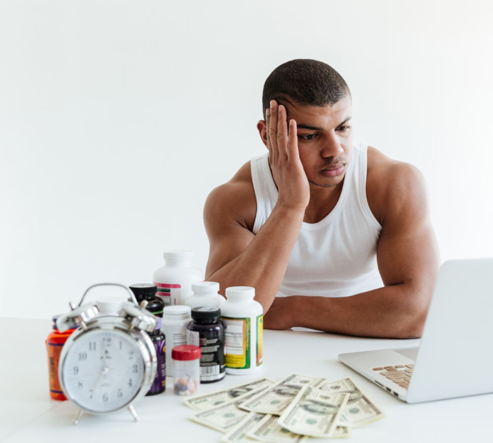 Man in tank top looks stressed at a laptop with supplements and money.