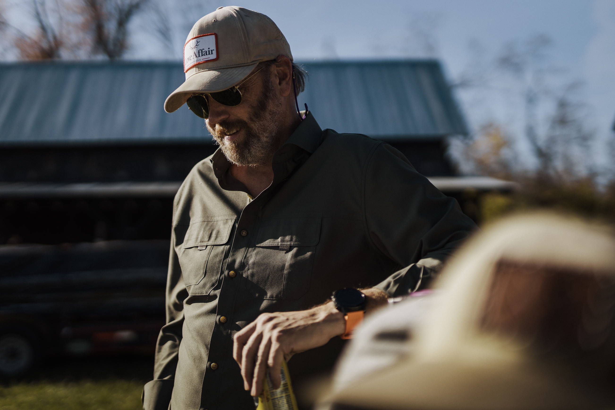 A man with a beard wearing the olive tulu custom fishing shirt, and hat, and sunglasses.