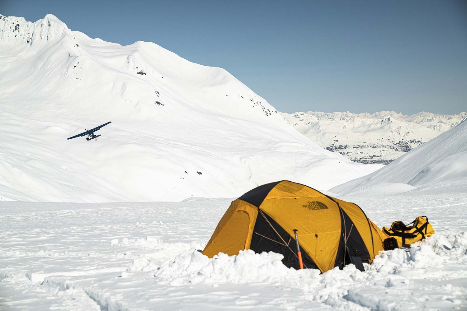 Yellow tent on snow-covered mountain landscape with airplane in the background.