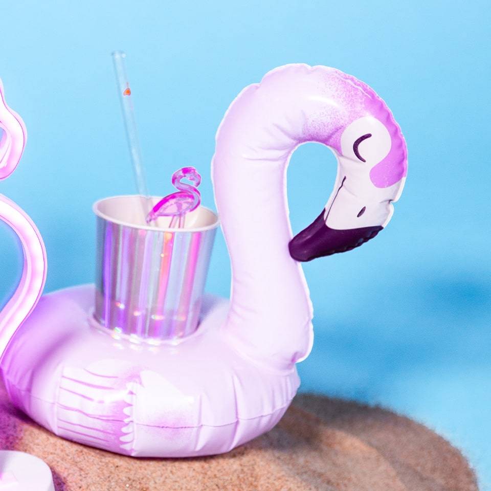 Inflatable flamingo drink holder on a sandy surface holding a metallic cup with a flamingo straw.