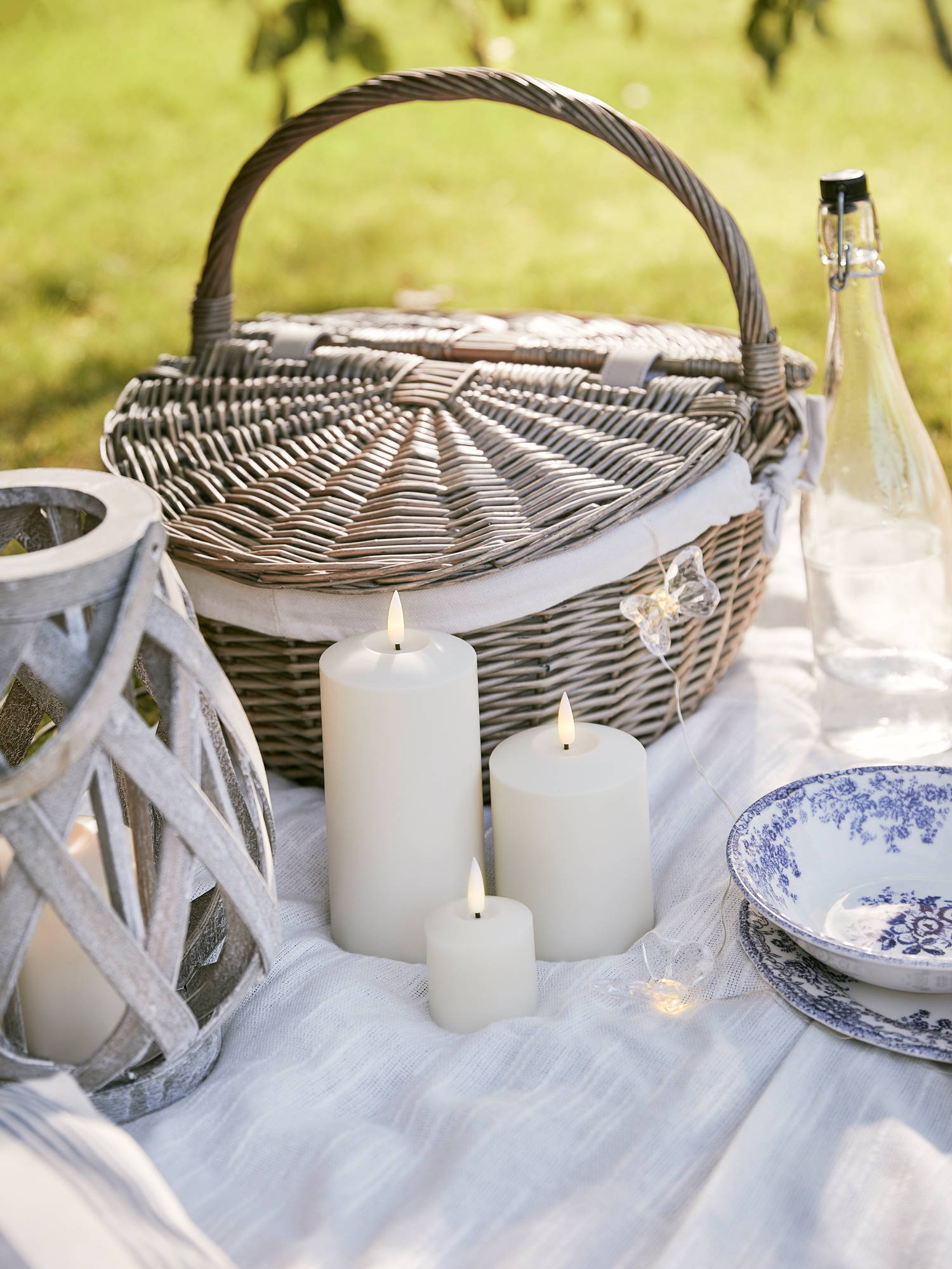 Outdoor TruGlow candles illuminated next to picnic basket on picnic blanket with butterfly micro lights and garden lantern