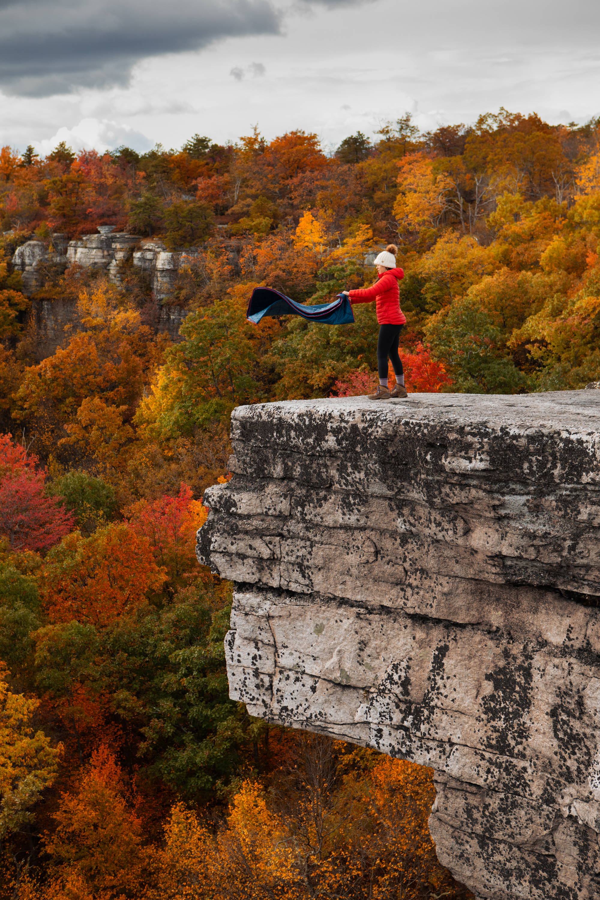 Woman shaking Rumpl blanket on cliff overlooking autumn landscape