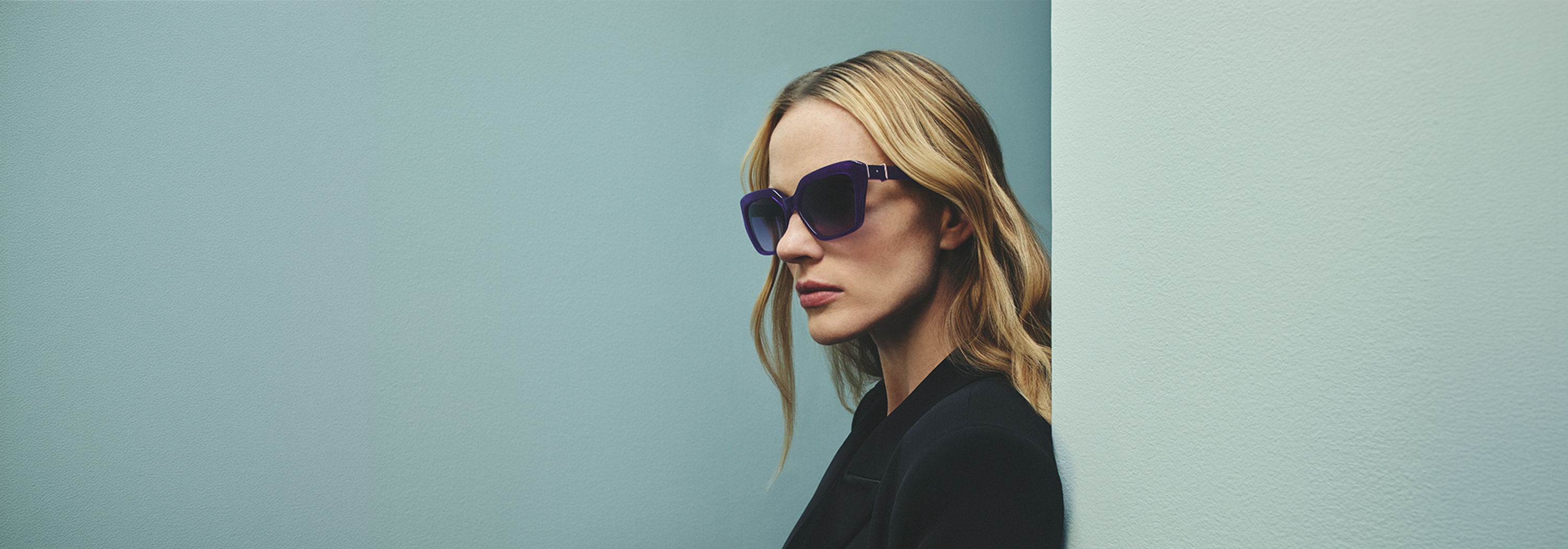 A woman in Robert Marc sunglasses standing against a light blue wall.