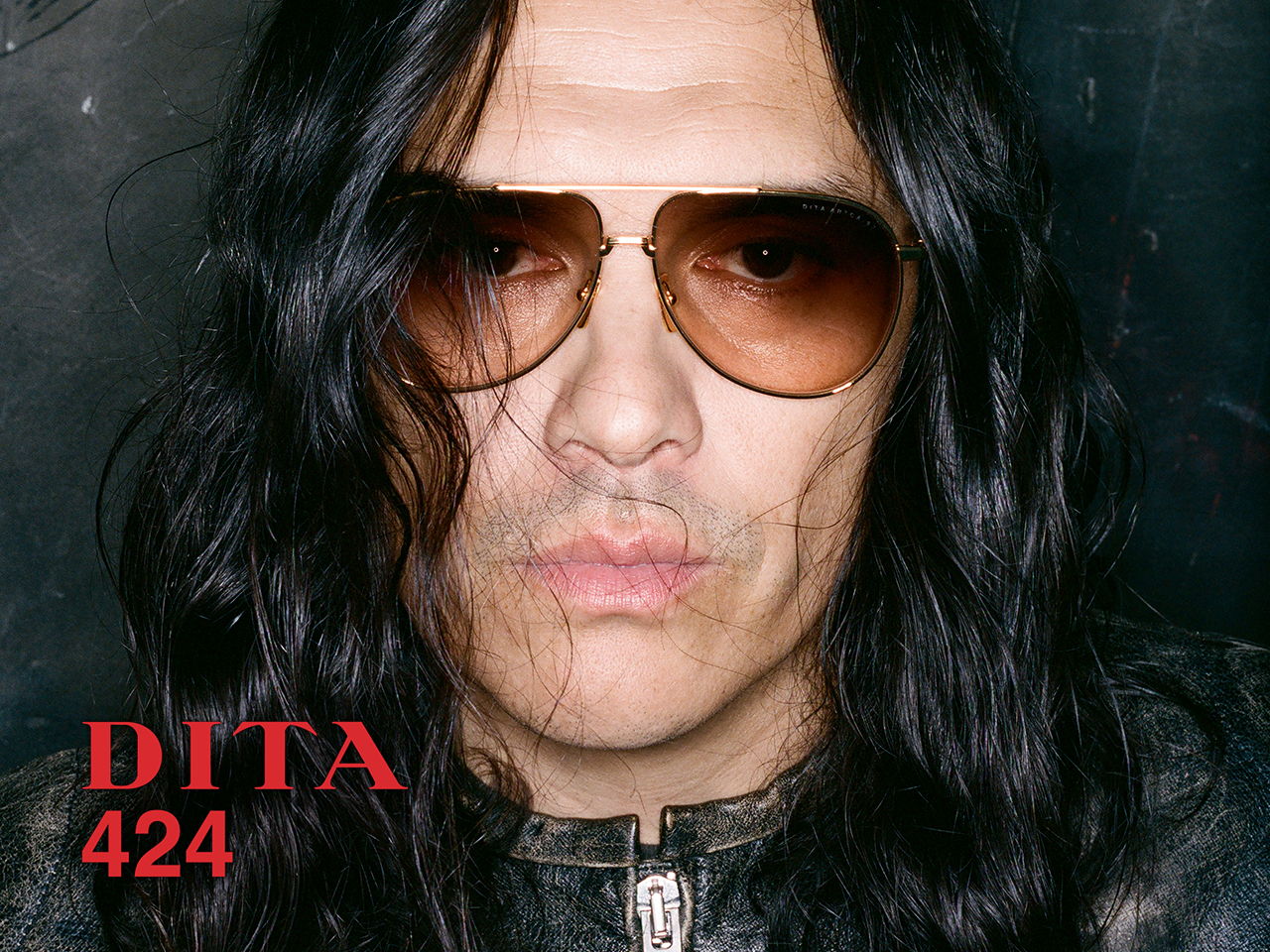 Person with long hair wearing sunglasses against a dark background.
