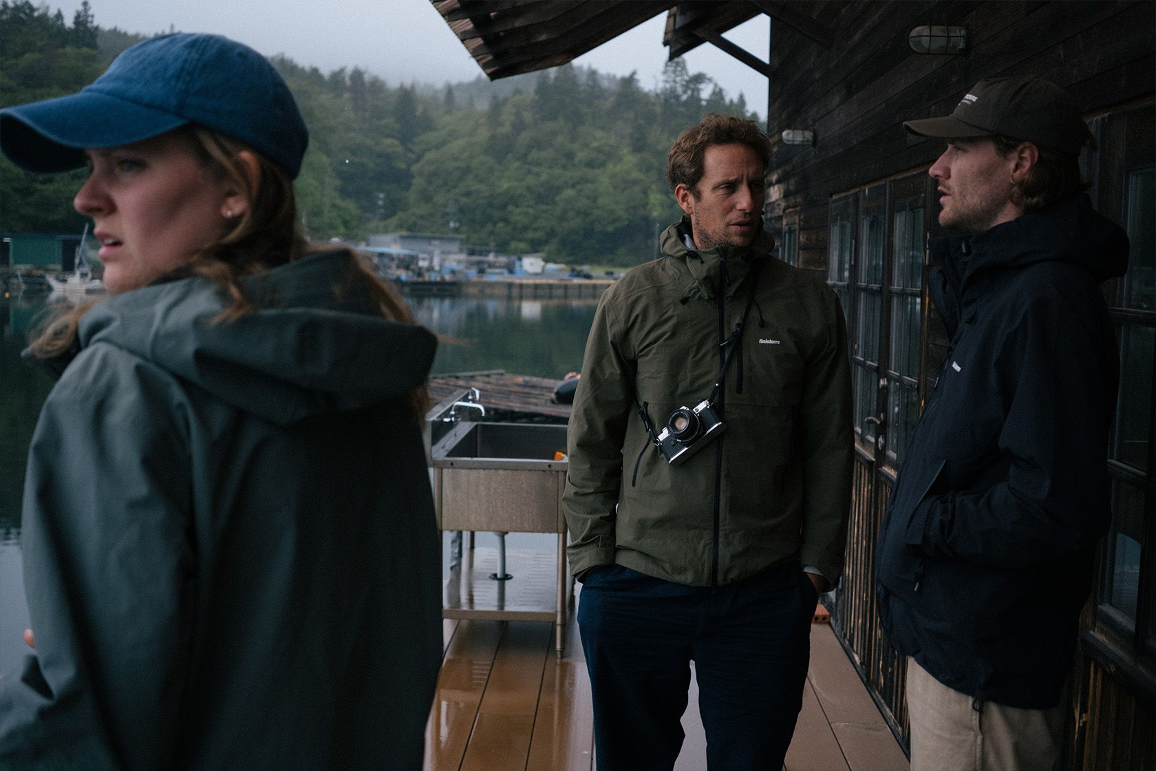 Three people stand on a wooden deck in a rainy setting.