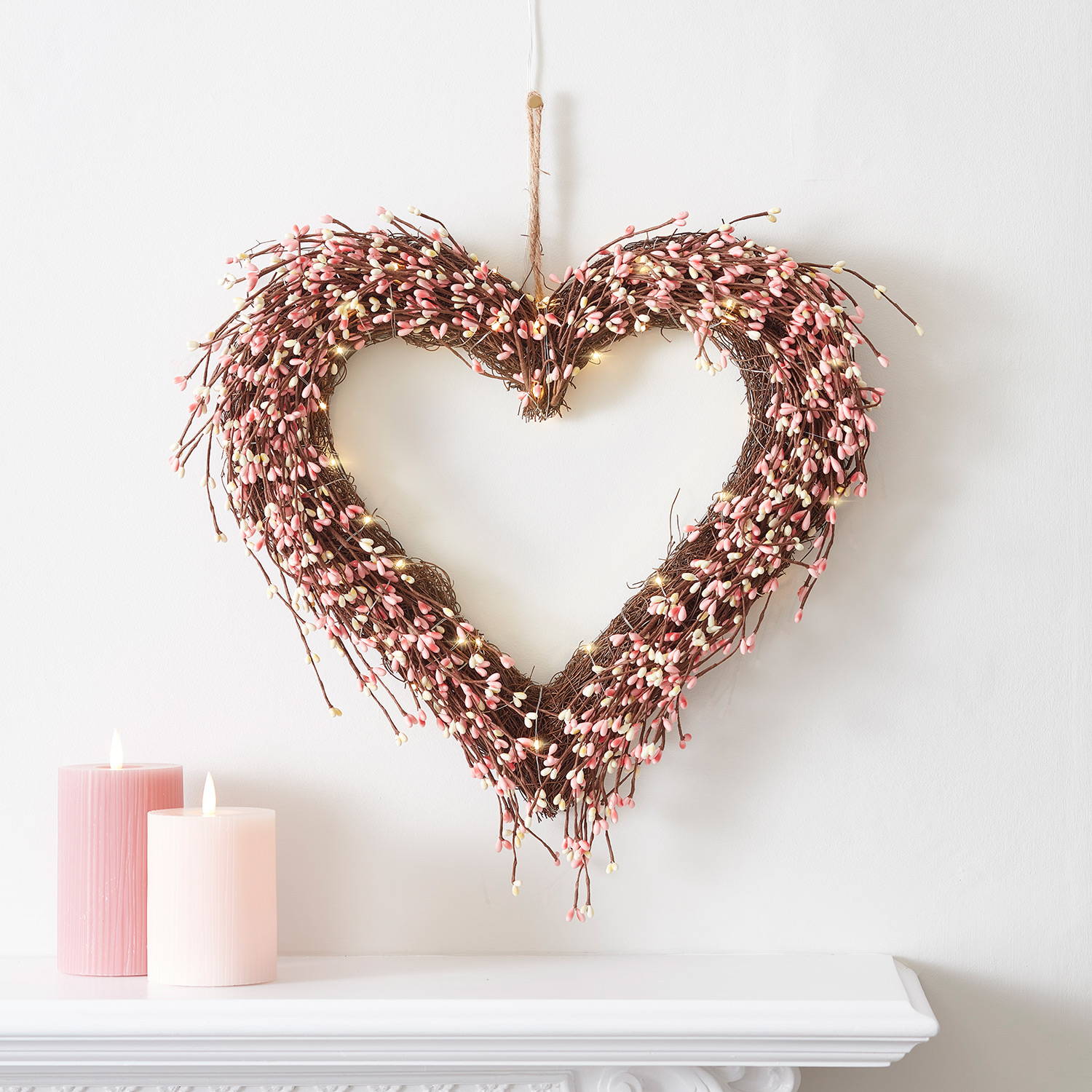 Pink berry heart wreath with warm white micro-lights on a white wall with LED candles on a mantlepiece.