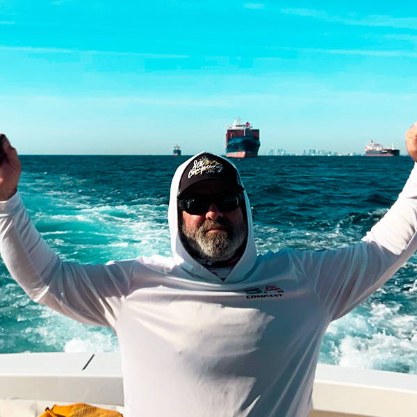 Jason Gascoyne in a boat, with his hands up in the air, wearing a SA Company hooded performance shirt, sunglasses, and a hat.