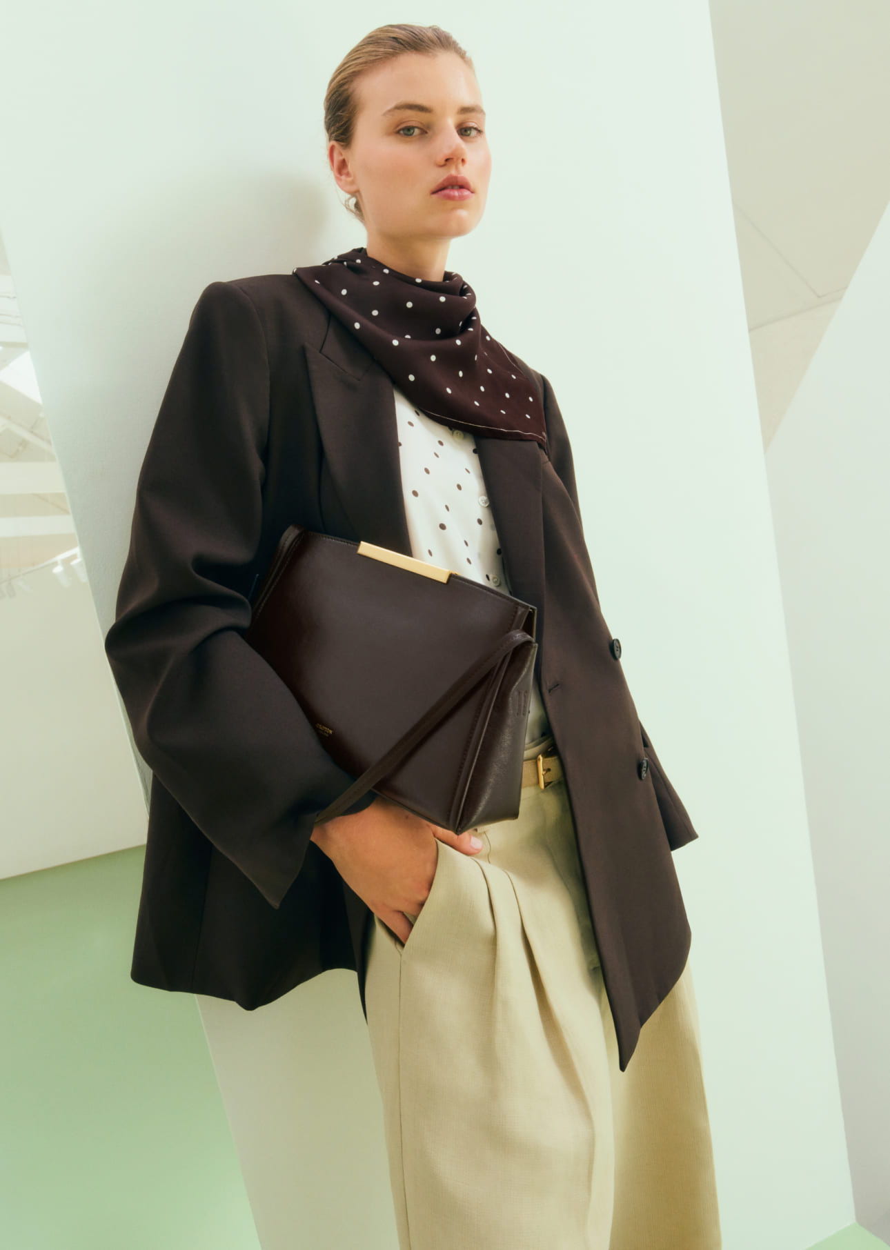 Woman in a stylish outfit holding a clutch, standing against a minimal backdrop.