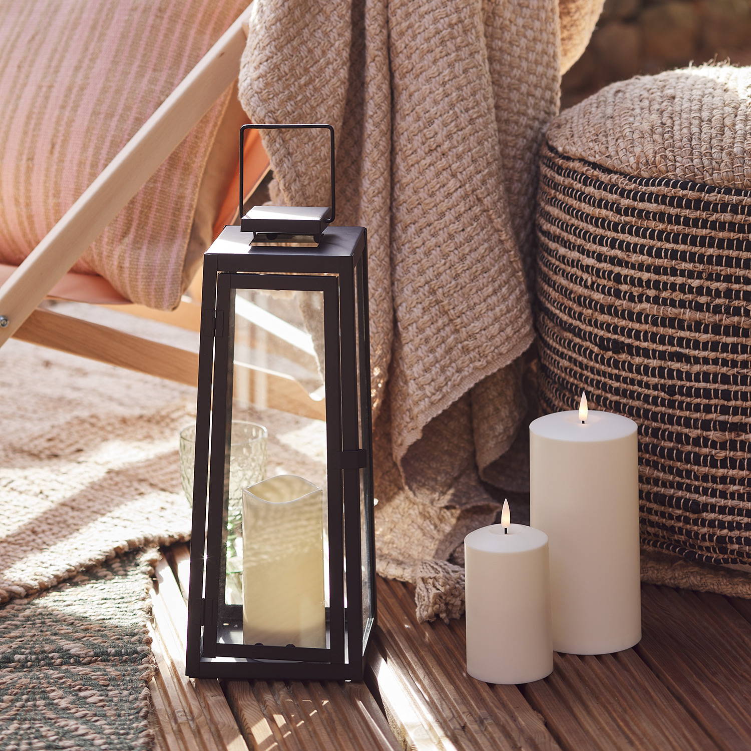 An outdoor black lantern and candles next to a comfy stool and blanket.