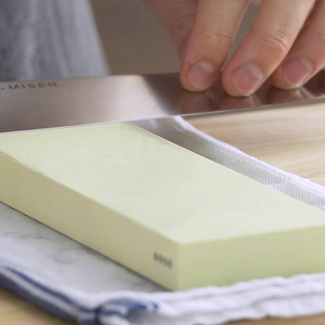 A chef using a cream-colored 6000-grit Misen Sharpening Stone to finish sharpening a Misen Chef’s Knife.