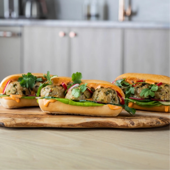 Three sandwiches with meatballs, vegetables, and sauce on a wooden board.