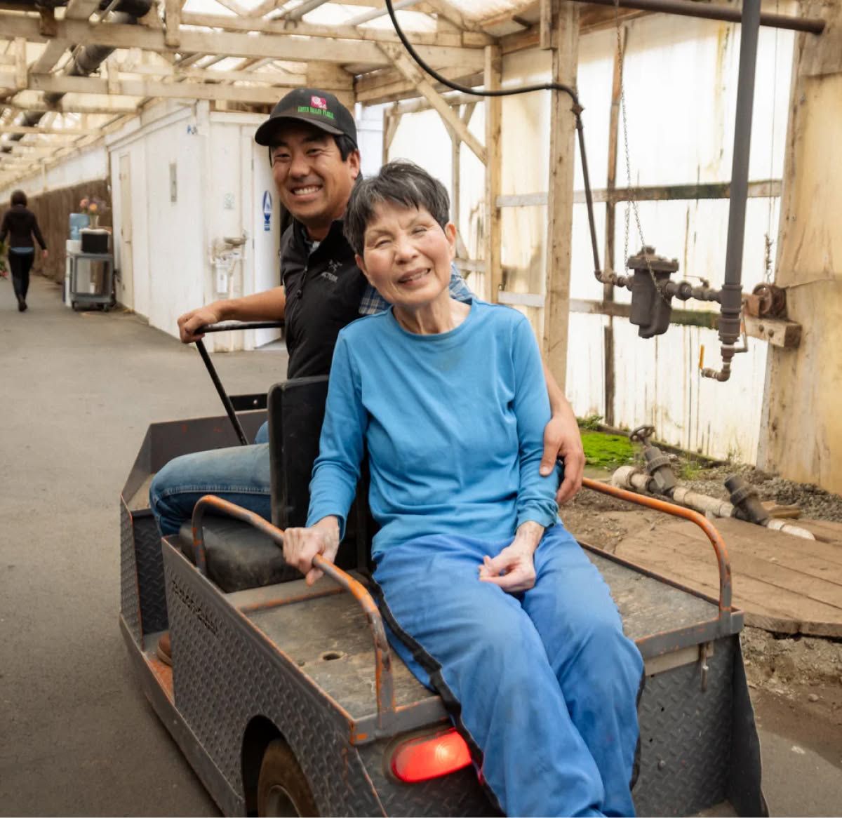 Two people smiling while sitting on a small vehicle in a greenhouse.