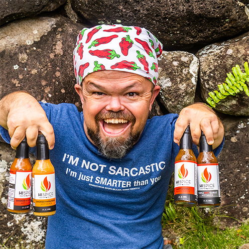 Man holding hot sauce bottles, wearing a humorous shirt and bandana.