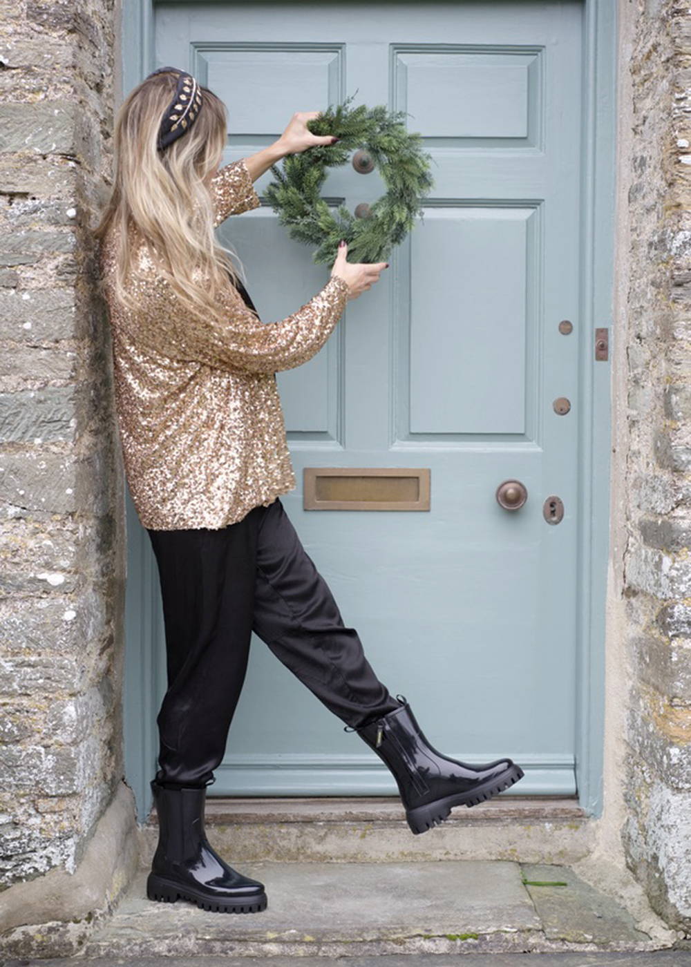 A model wearing a gold sparkly jacket, black trousers and black boots hanging a wreath 