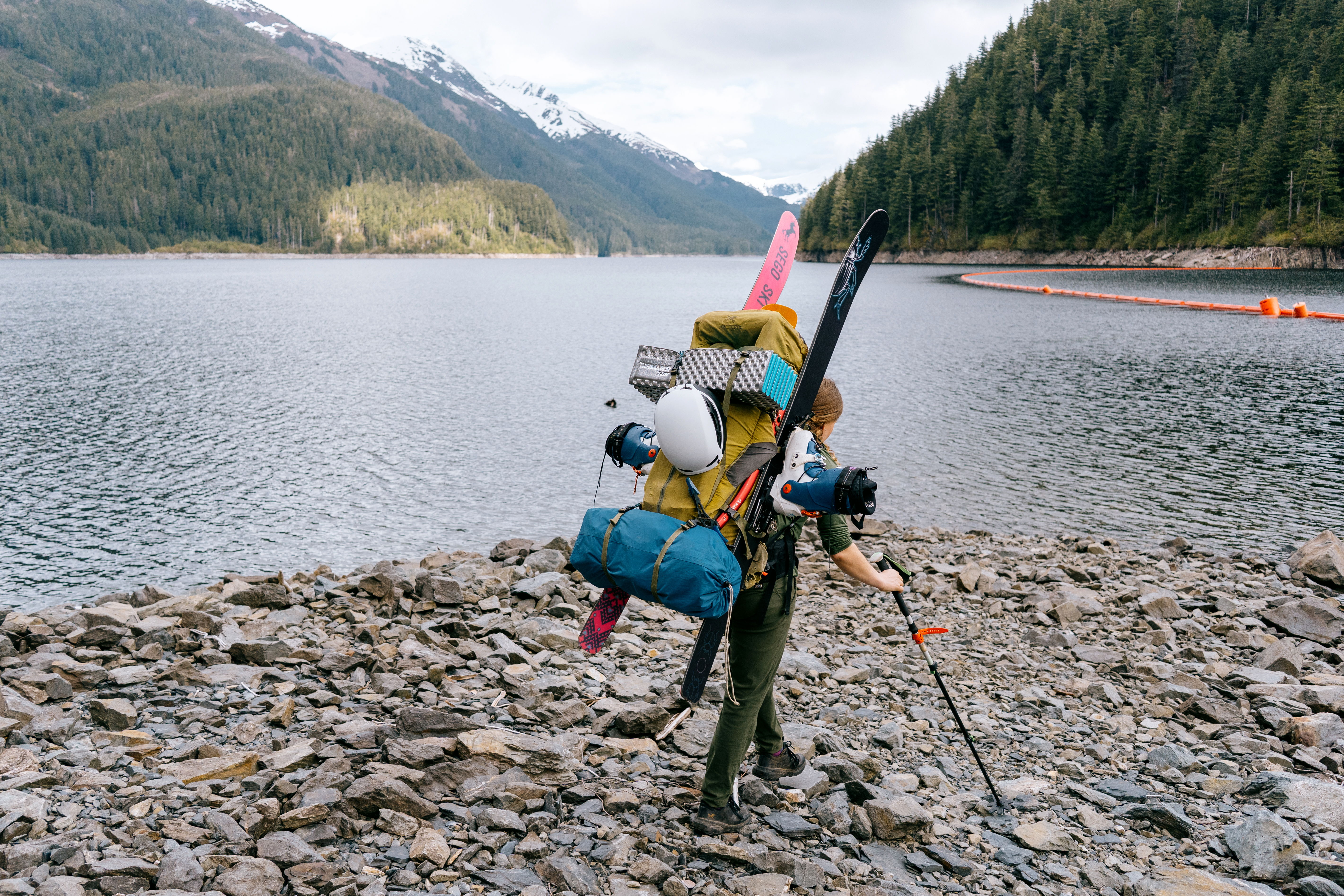 Person with a backpack standing by a lake surrounded by mountains.