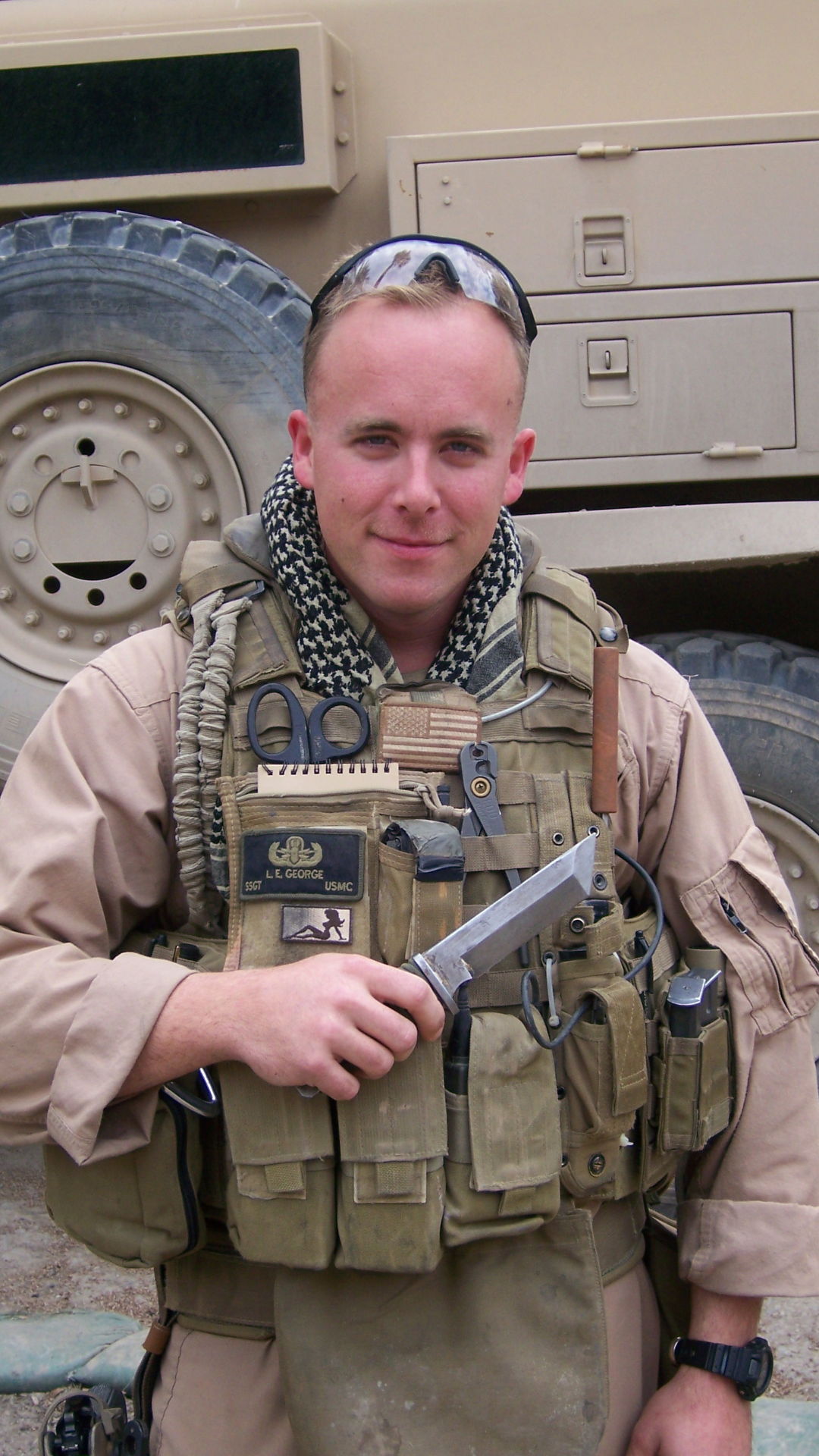 Soldier in tactical gear holding a handgun, standing near military vehicle.
