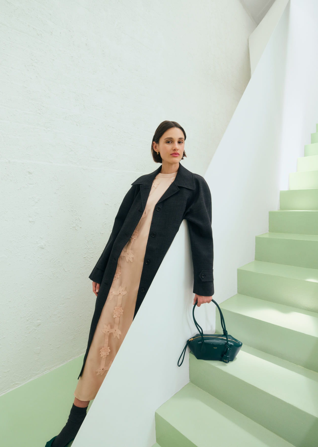 Woman in a black coat poses on light green stairs, holding a handbag.