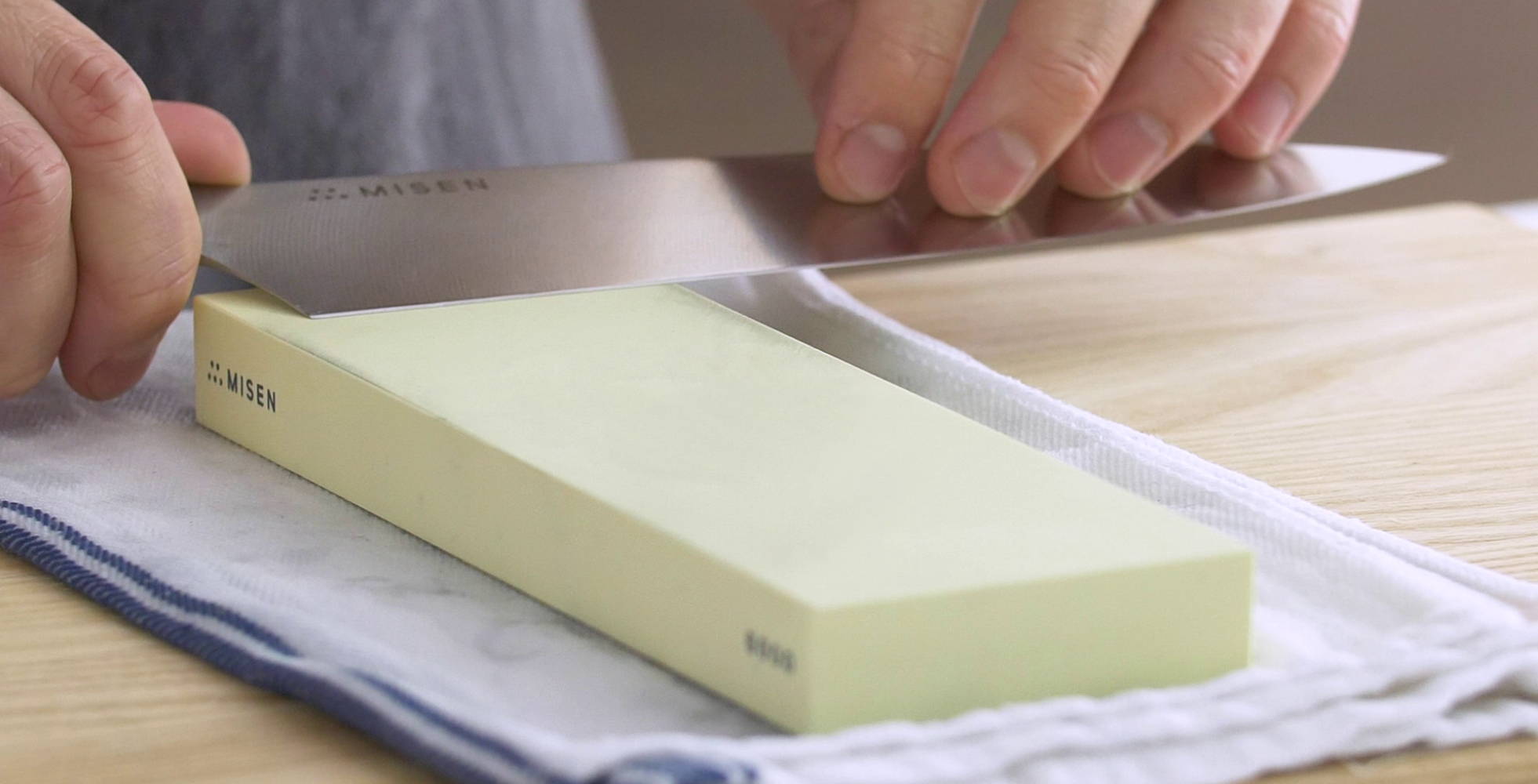A chef using a cream-colored 6000-grit Misen Sharpening Stone to finish sharpening a Misen Chef’s Knife.