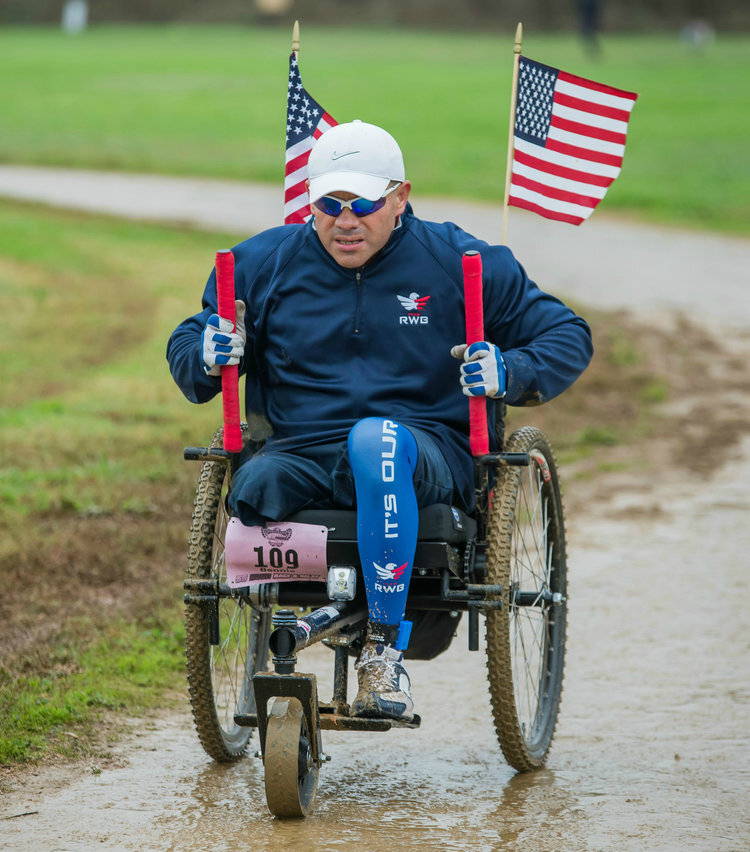 Person with lower limb loss pushes levers of GRIT Freedom Chair on muddy dirt path with American flags flying off back of wheelchair
