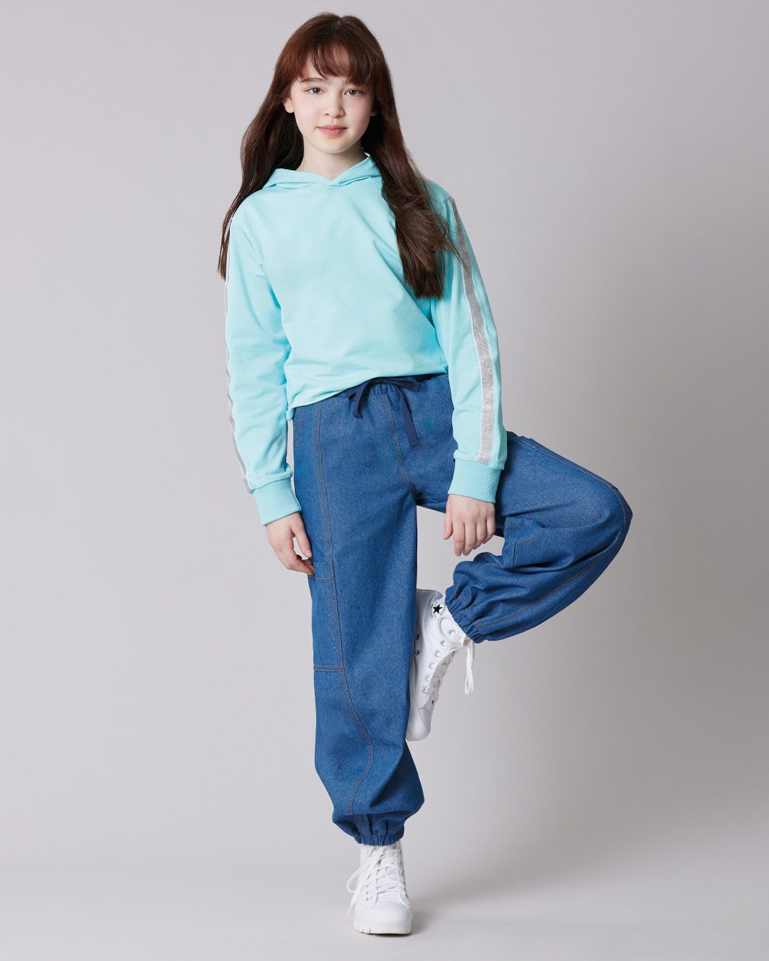 Girl in a turquoise hoodie and blue pants posing against a gray background.