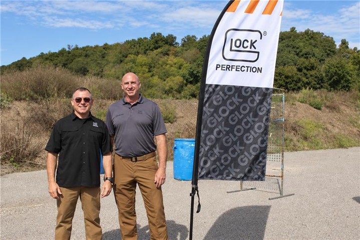 Two men stand beside a flag at an outdoor event.