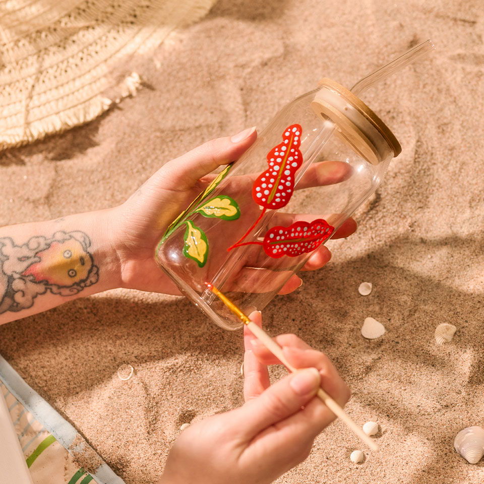 Person holding a decorated glass with a straw on sandy surface.