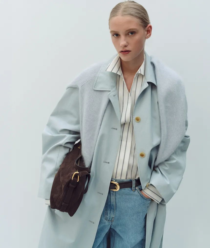 Model in light blue coat holding a brown handbag against a plain background.