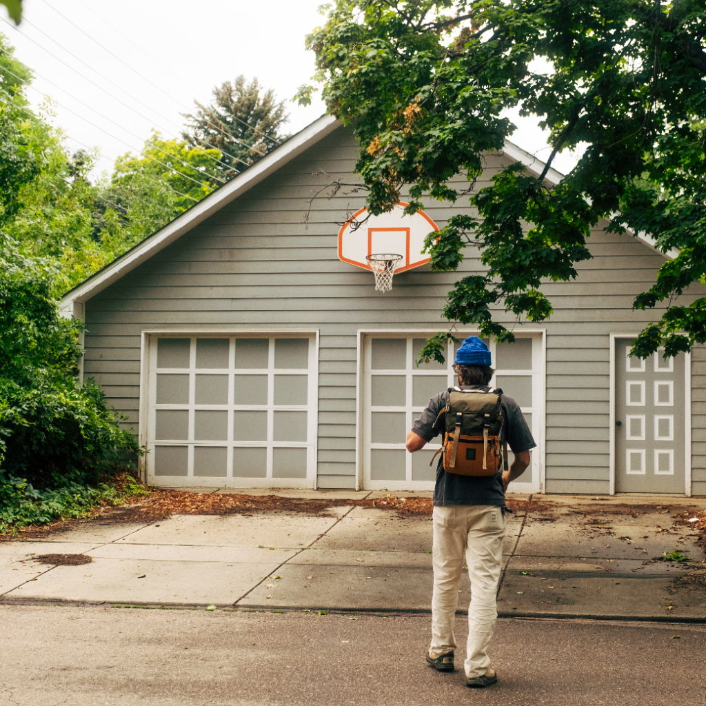 Person walking towards a garage with a basketball hoop.