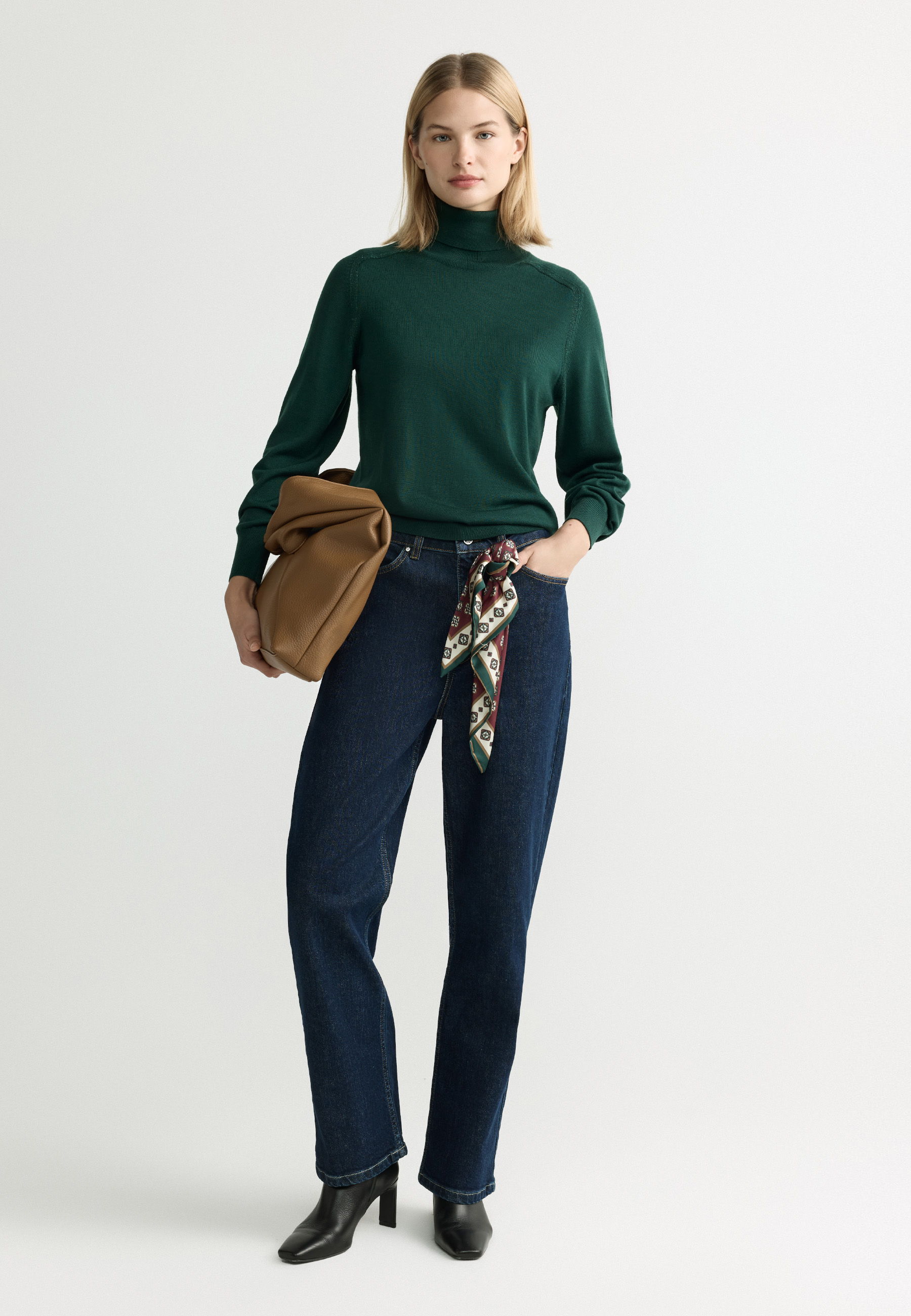Woman in green sweater and dark jeans, holding a brown bag.