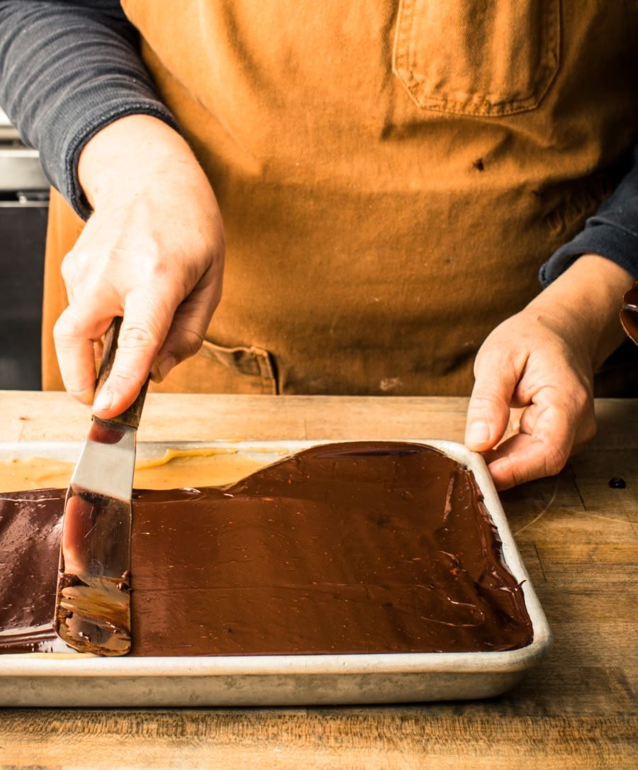 Person spreading chocolate frosting in a baking dish on a wooden surface.
