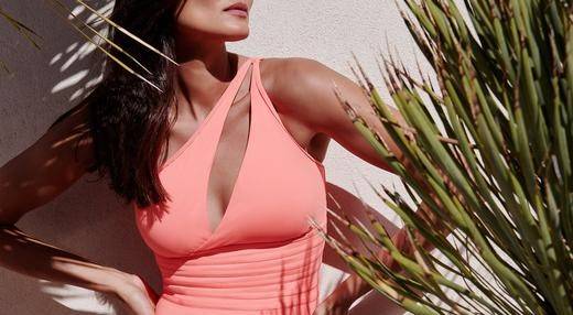 Model wearing a pink one shoulder swimsuit with a plant next to her.