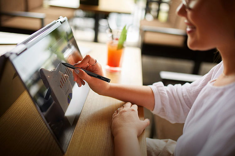 Smiling woman using a stylus on a 2-in-1 laptop showing charts, seated at a wooden table 