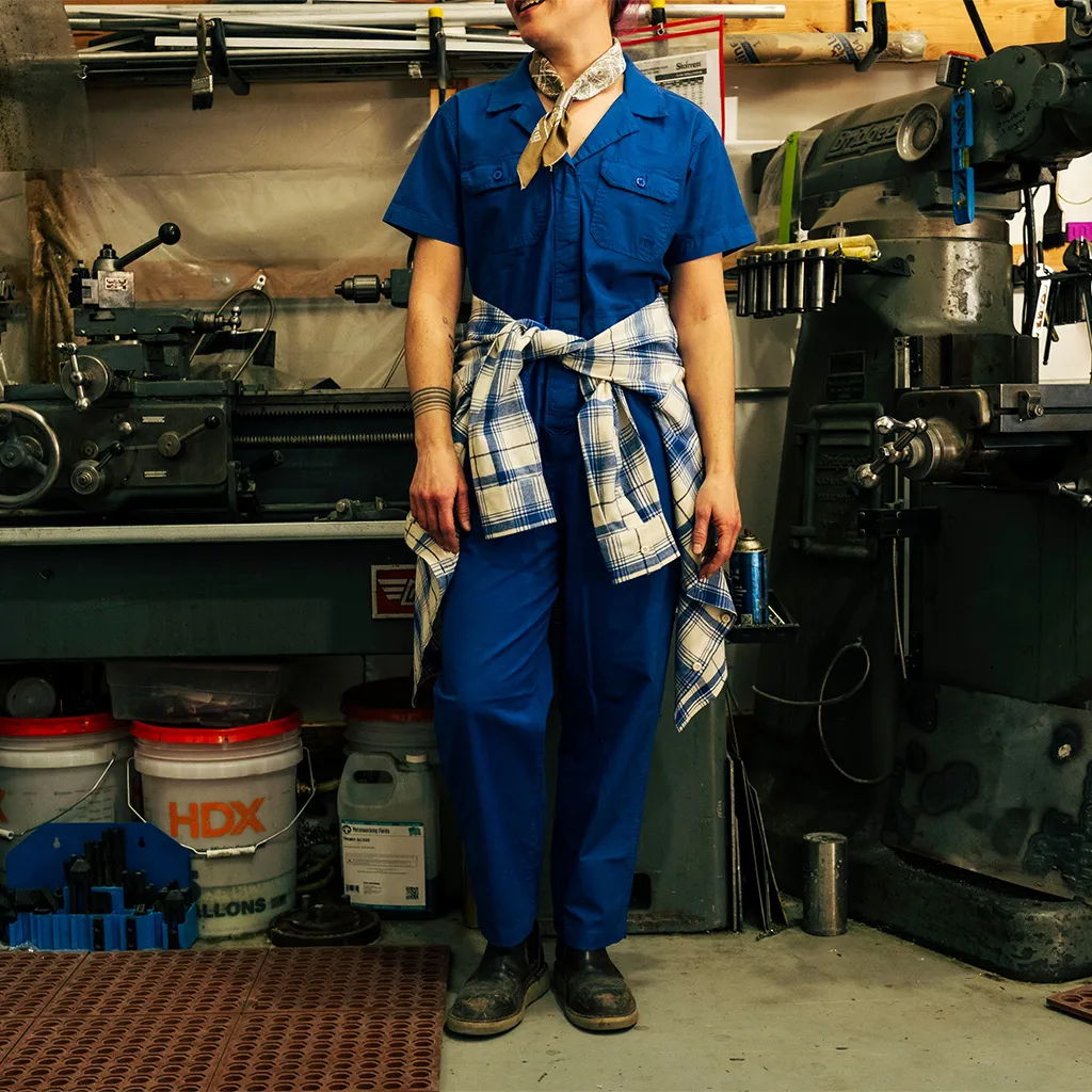 Person in blue coveralls standing in a workshop with tools and equipment.