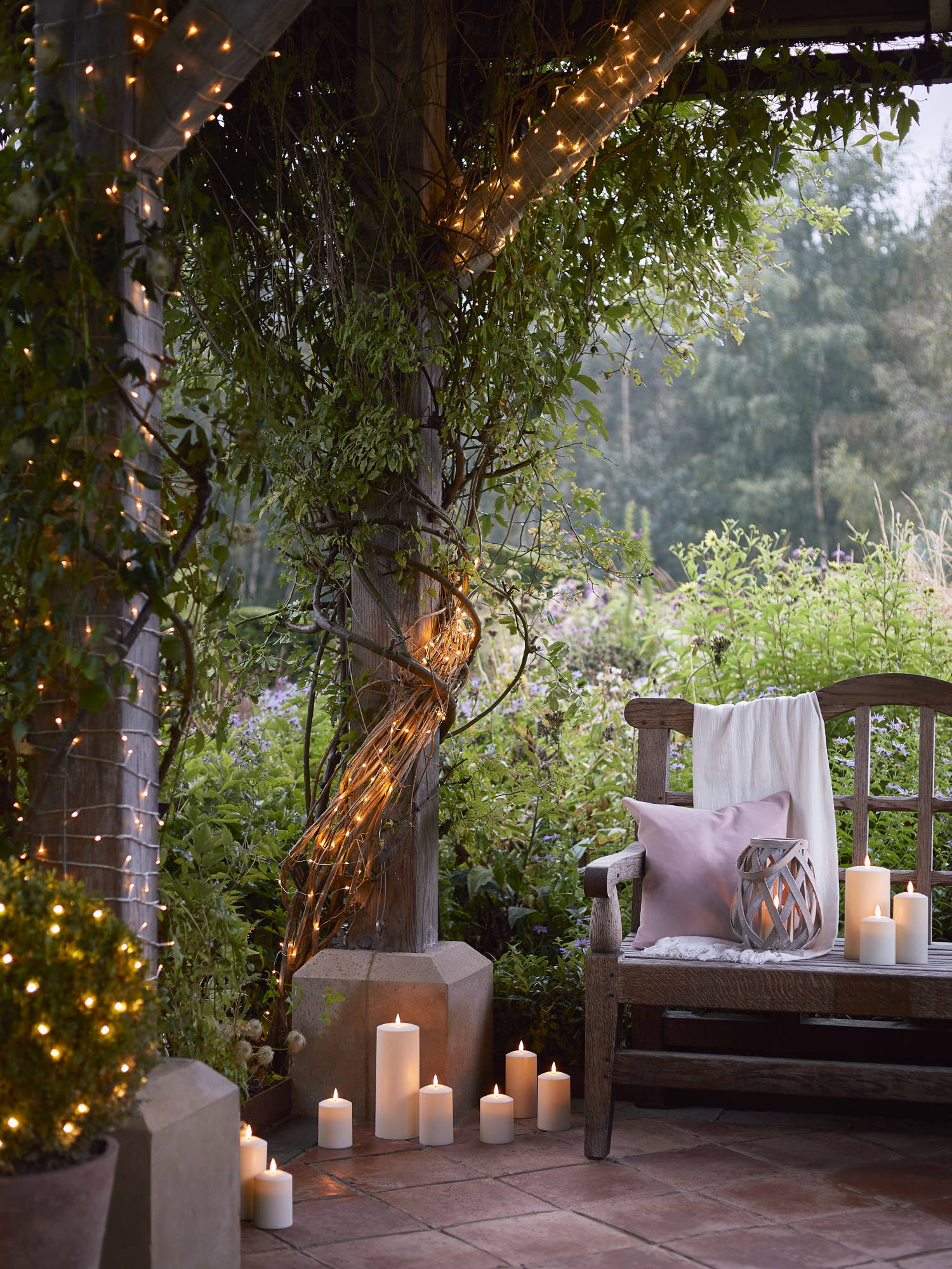 Outdoor spring summer setting with garden lantern sat on bench with TruGlow candles scattered on terrace area and fairy lights illuminated