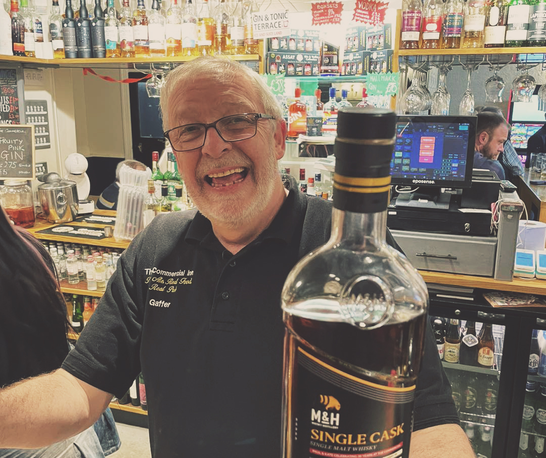 Smiling bartender holding a bottle of whiskey in a lively bar setting.