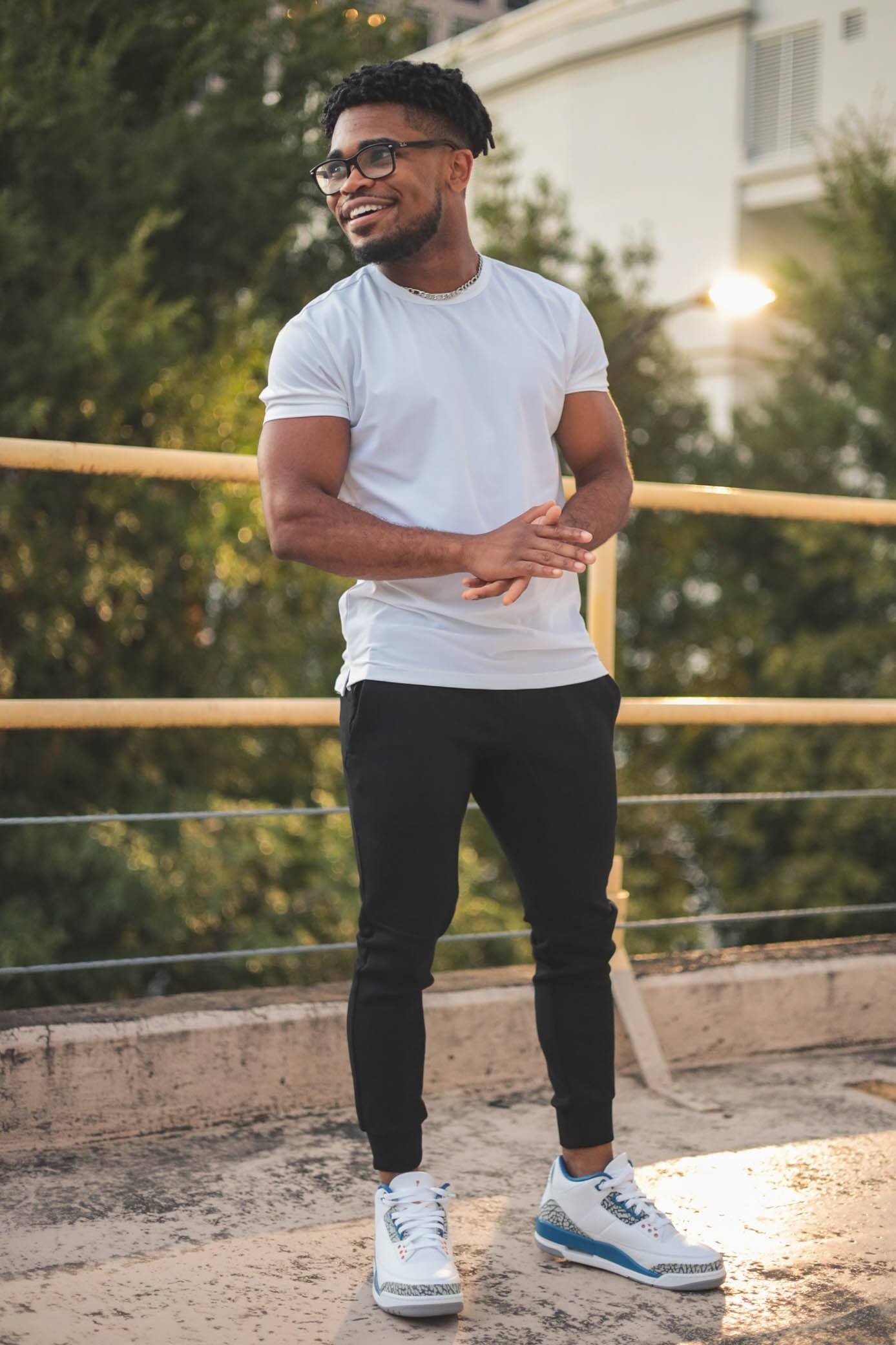 Man standing and smiling with hands together wearing jordan sneakers, a white t shirt, and black flex joggers from under510.com